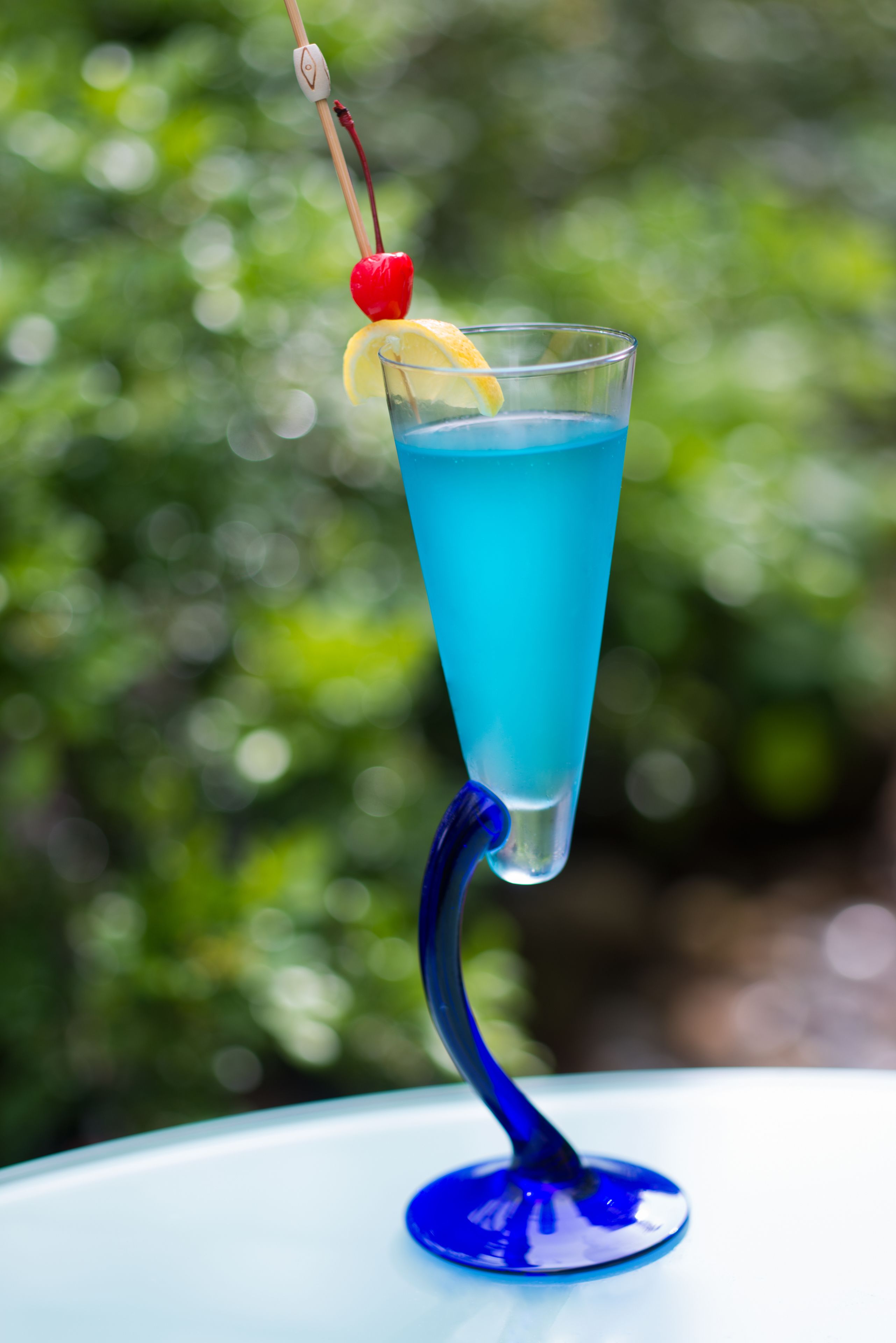 A blue cocktail with a lemon slice and cherry garnish on a blue stemmed glass.