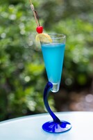 A blue cocktail with a lemon slice and cherry garnish on a blue stemmed glass.