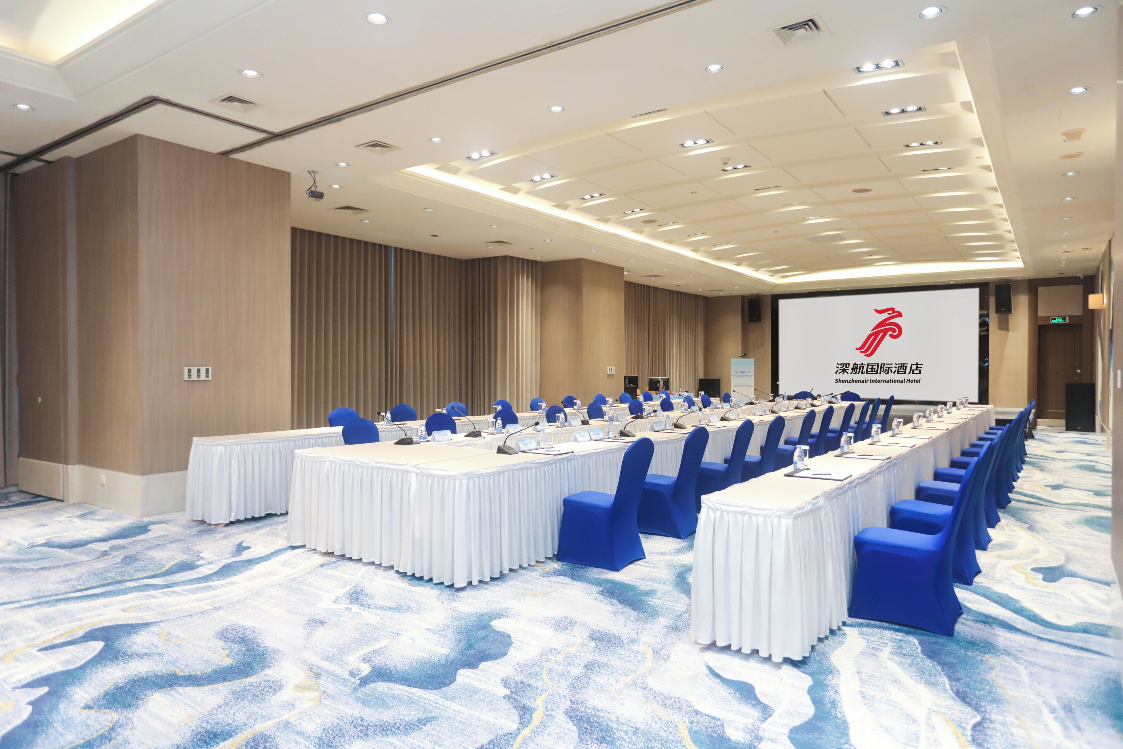 Un salón de conferencias con una larga mesa, sillas azules y una pantalla proyectora que muestra el logotipo de un hotel.