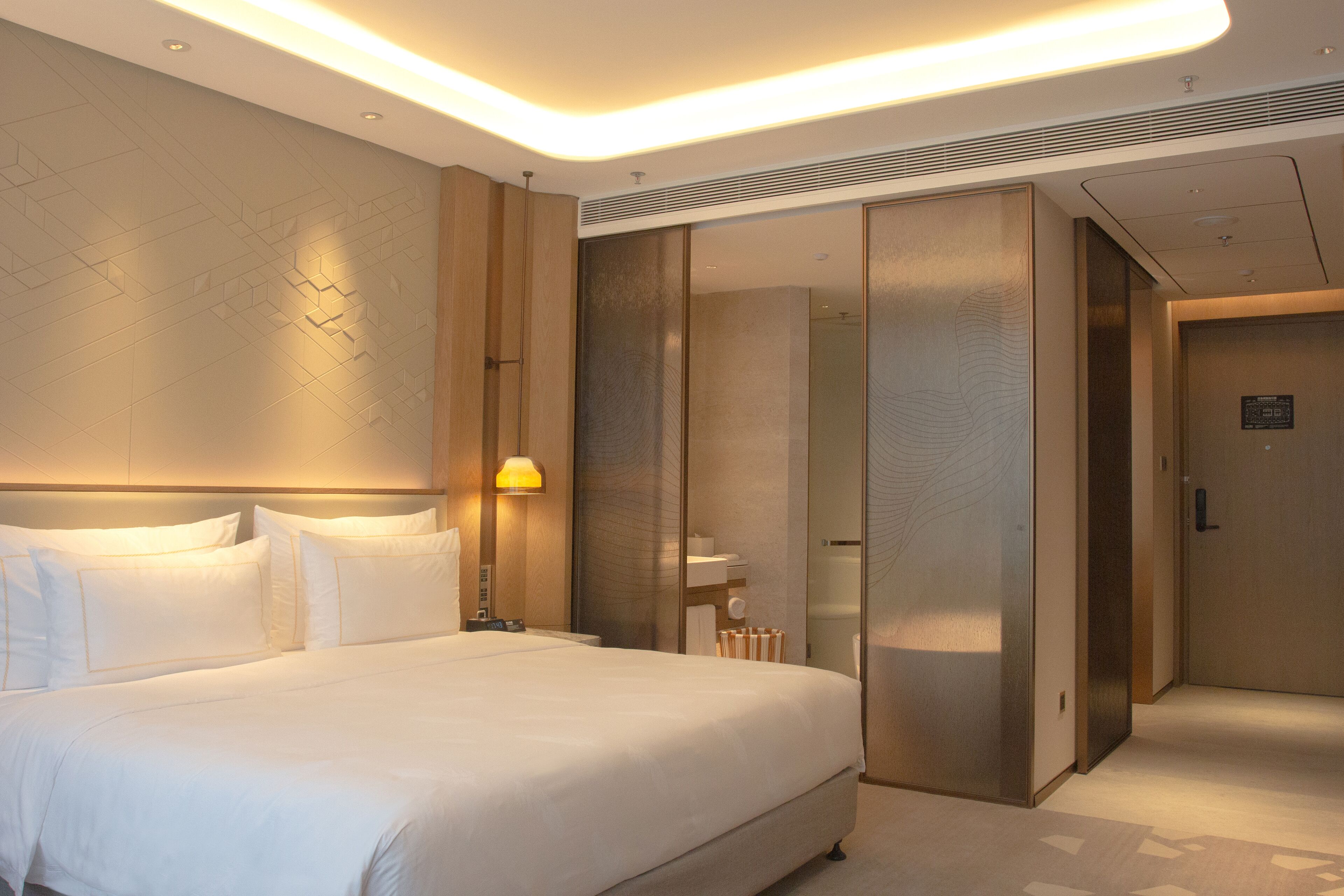 A modern hotel room with a large bed, a bedside table, a lamp, and a sliding door leading to a bathroom.
