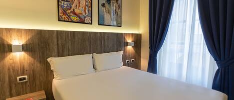 A hotel room with a large bed, two wall-mounted lights, a wooden headboard, and two framed artworks above the bed.