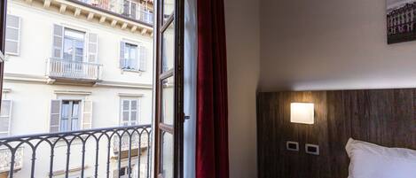 A hotel room with a bed, a nightstand, a wall-mounted light, and a window with a view of a building.
