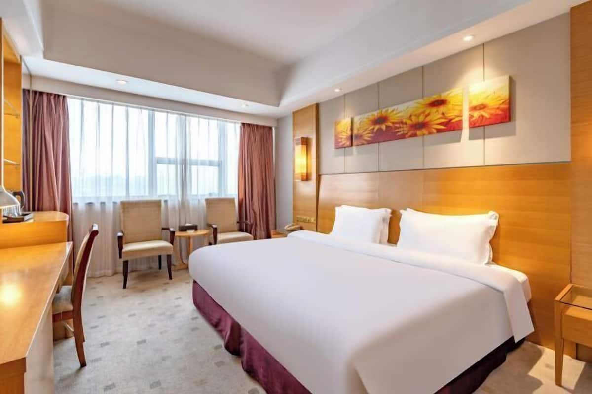 A hotel room with a large bed, a desk, two chairs, a window with curtains, and a wall with a floral painting.