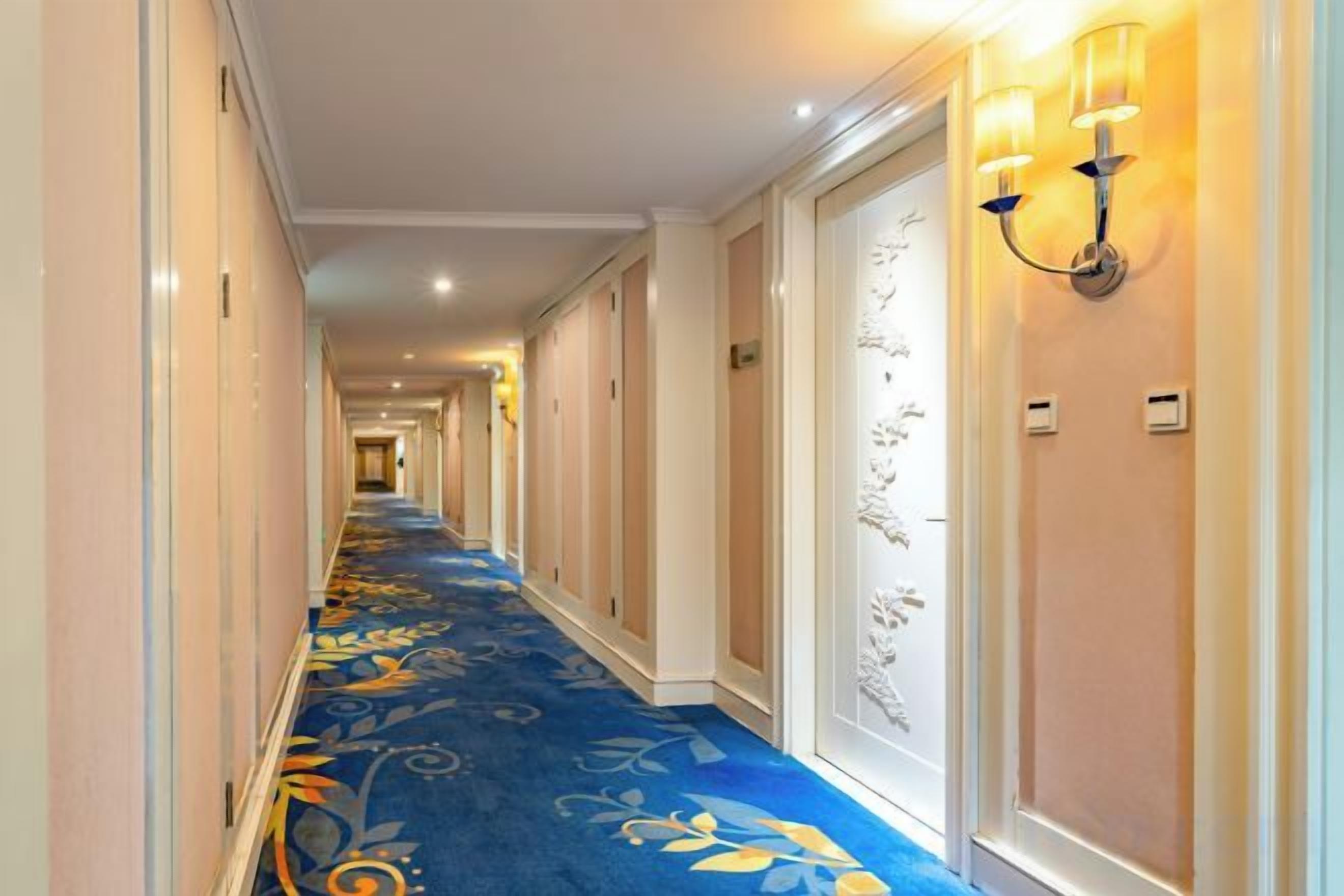 A hotel corridor with a floral carpet, wall sconces, and doors leading to rooms.