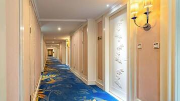 A hotel corridor with a floral carpet, wall sconces, and doors leading to rooms.