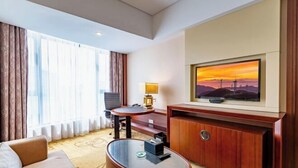 A hotel room with a sofa, a round glass coffee table, a TV on a wooden stand, a desk with a chair, and a large window with curtains.