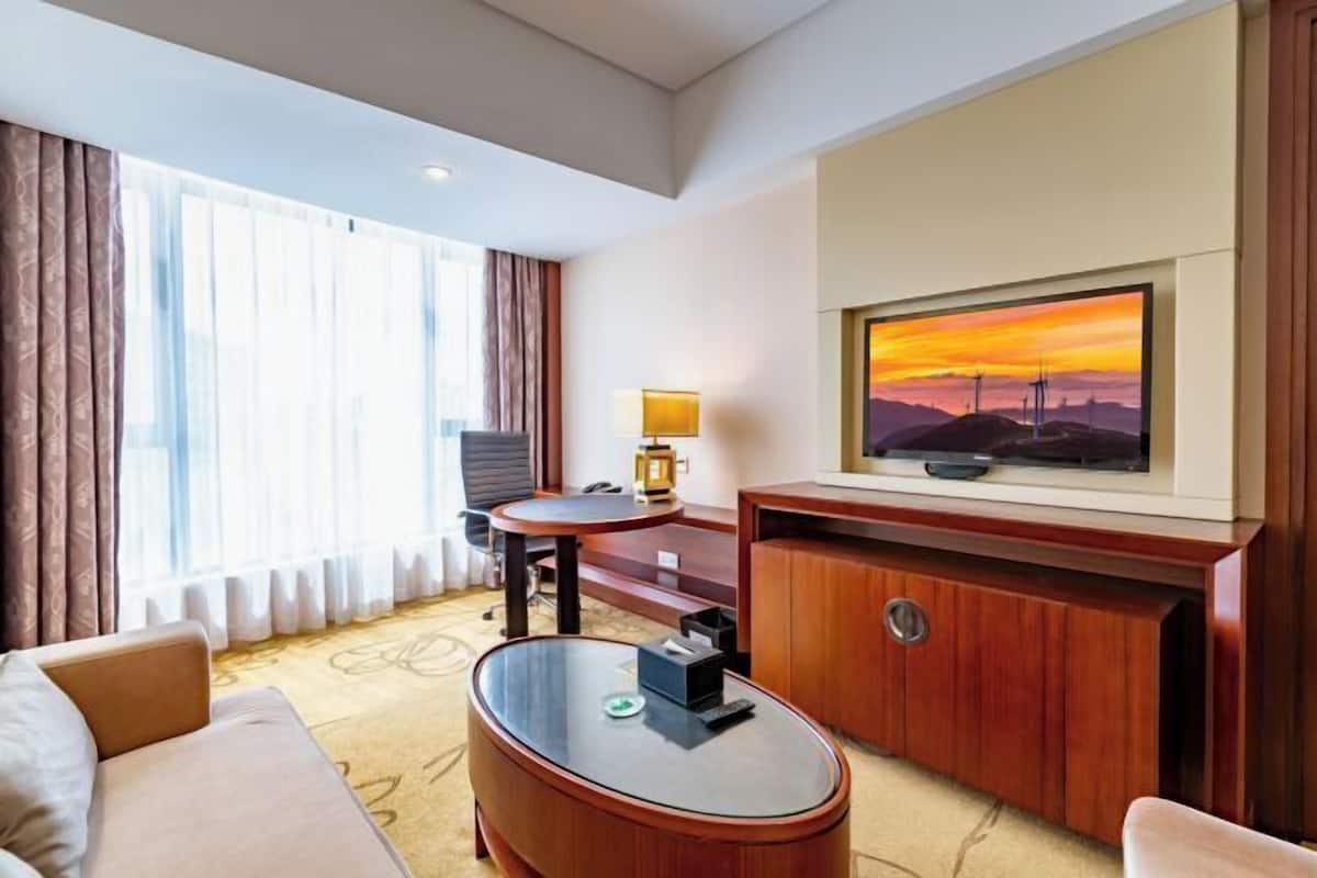 A hotel room with a sofa, a round glass coffee table, a TV on a wooden stand, a desk with a chair, and a large window with curtains.