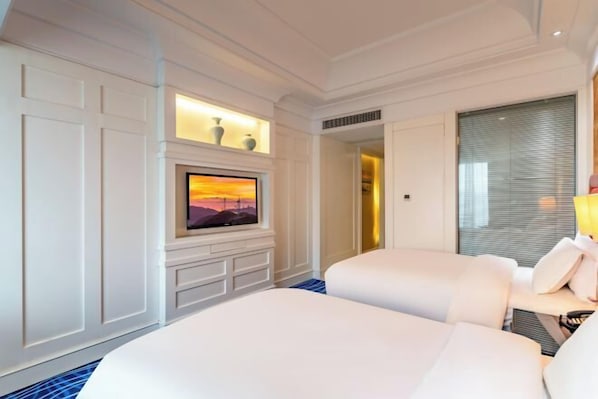A hotel room with two beds, a TV mounted on the wall, a small table with a lamp, and a window with blinds.