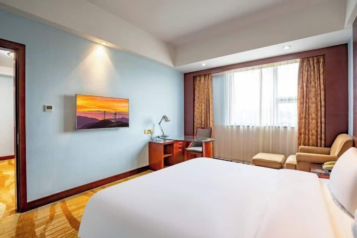 A hotel room with a large bed, a desk with a chair, a lamp, a painting, and a window with curtains.