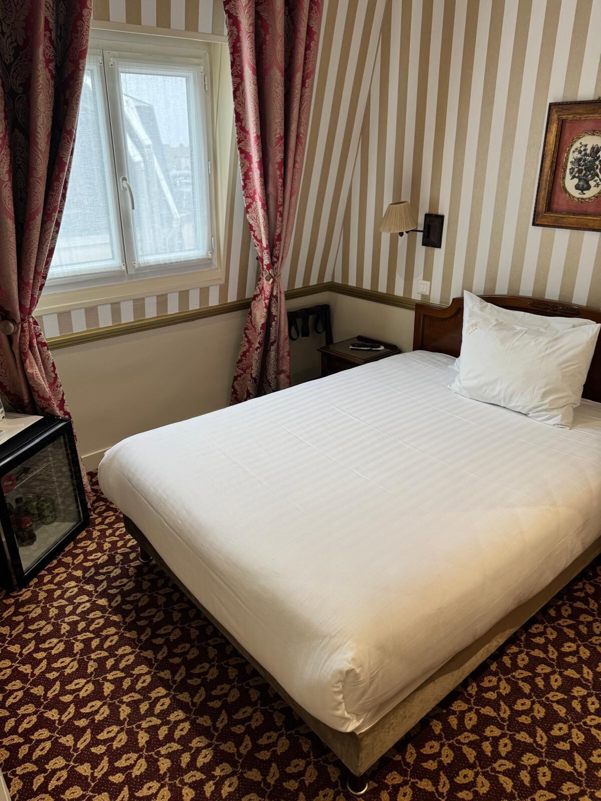 A hotel room with a bed, a nightstand, a lamp, a framed picture on the wall, and a window with curtains.