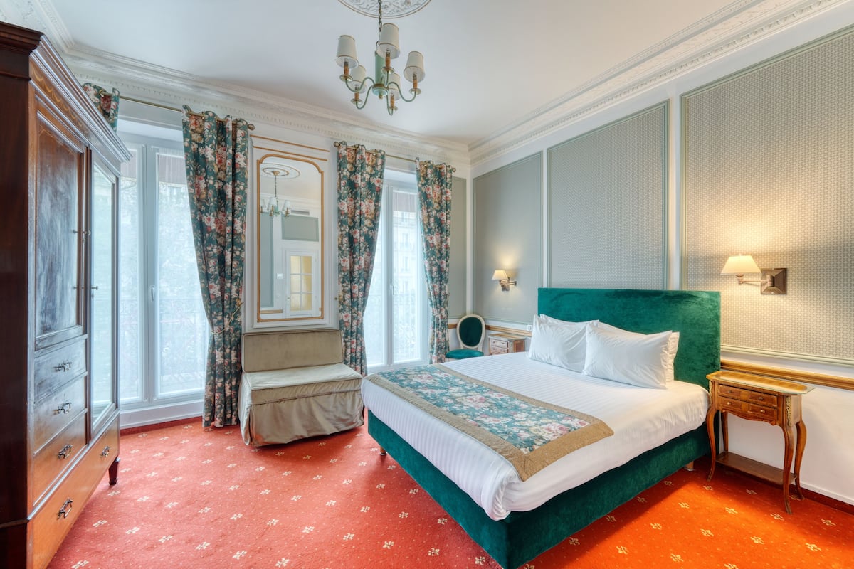 A hotel room with a large bed, a wooden dresser, a bench, and a window with curtains.