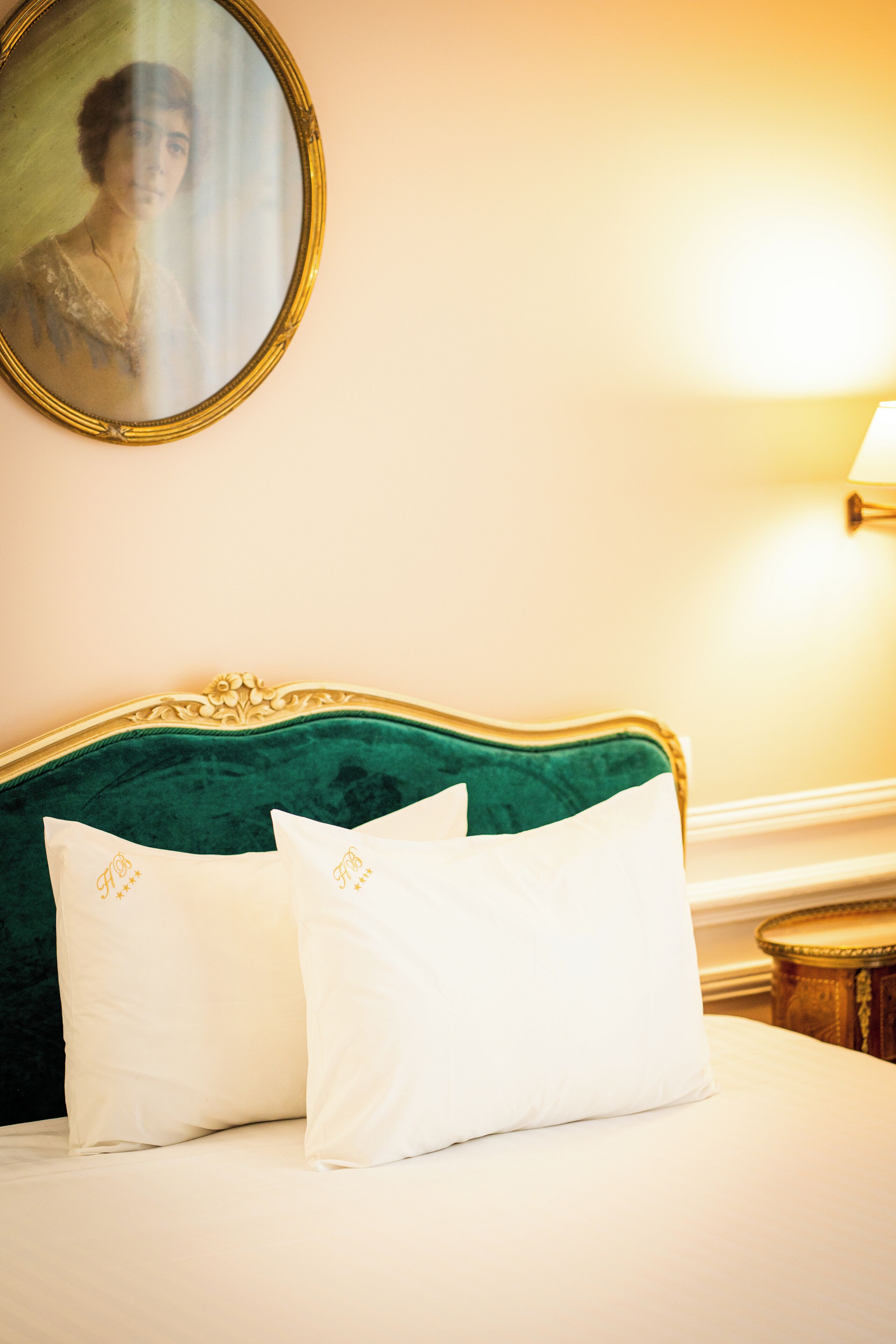 Un lit avec une tête de lit recouverte d’un tissu vert, deux oreillers blancs ornés de motifs brodés, et un miroir rond avec un cadre doré accroché au mur.