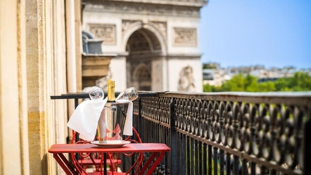 A balcony with a table set for two, a bottle of champagne, and a glass of wine. In the background, the Arc de Triomphe is visible.