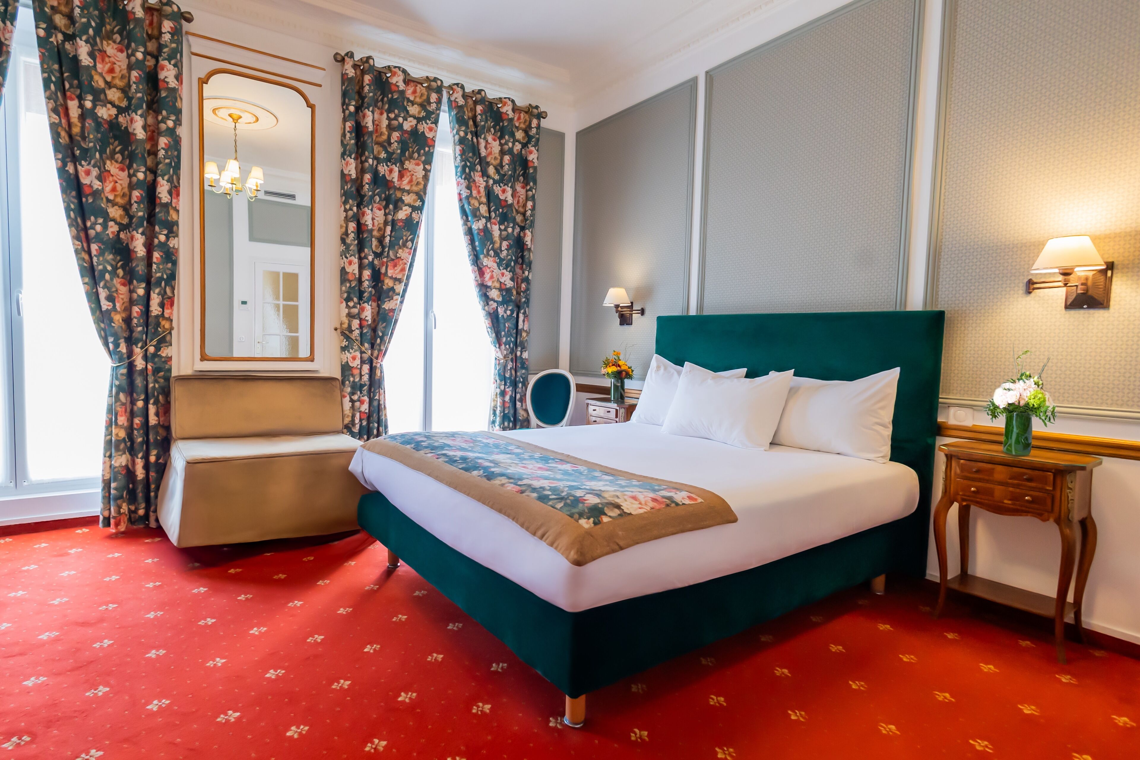 A hotel room with a large bed, a bedside table with a lamp, a mirror, and floral curtains.