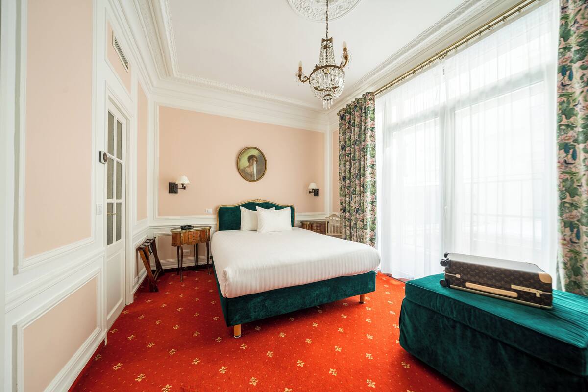 A bedroom with a large bed, a dresser, a chandelier, and a window with curtains.