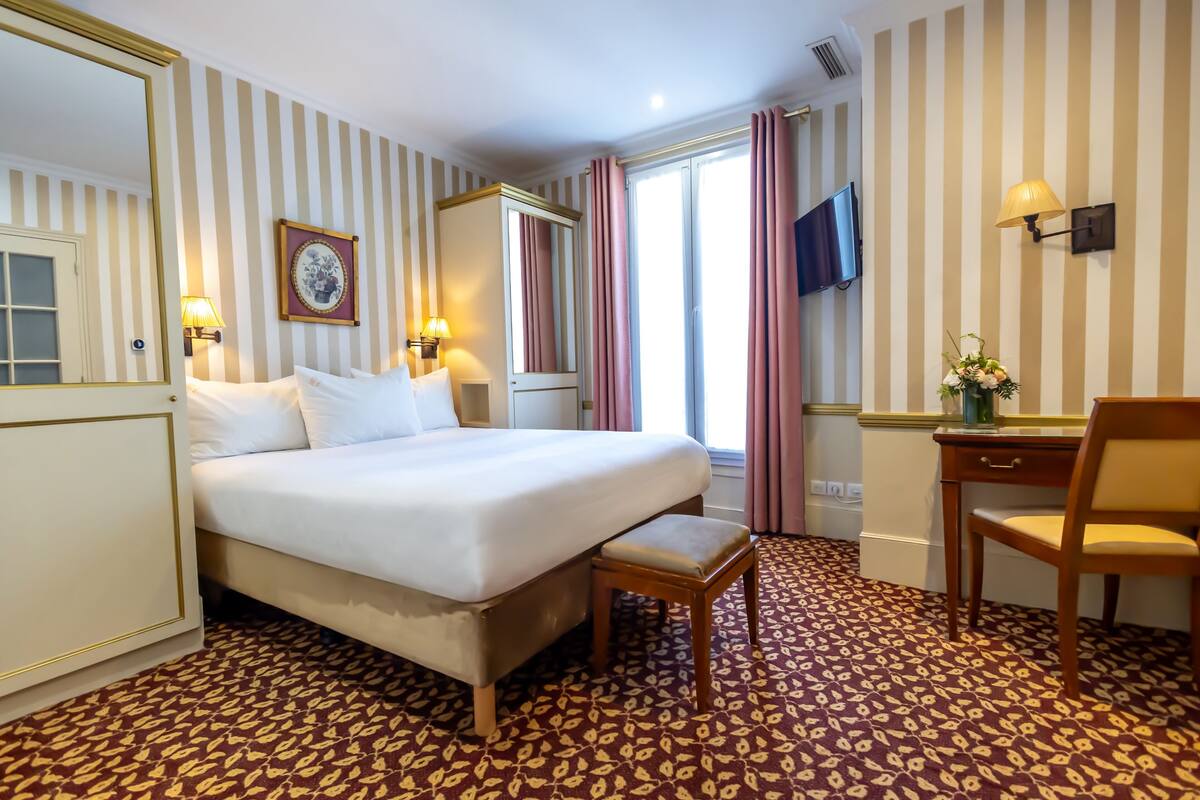 A hotel room with a large bed, a desk, a chair, a TV, and a window with curtains.