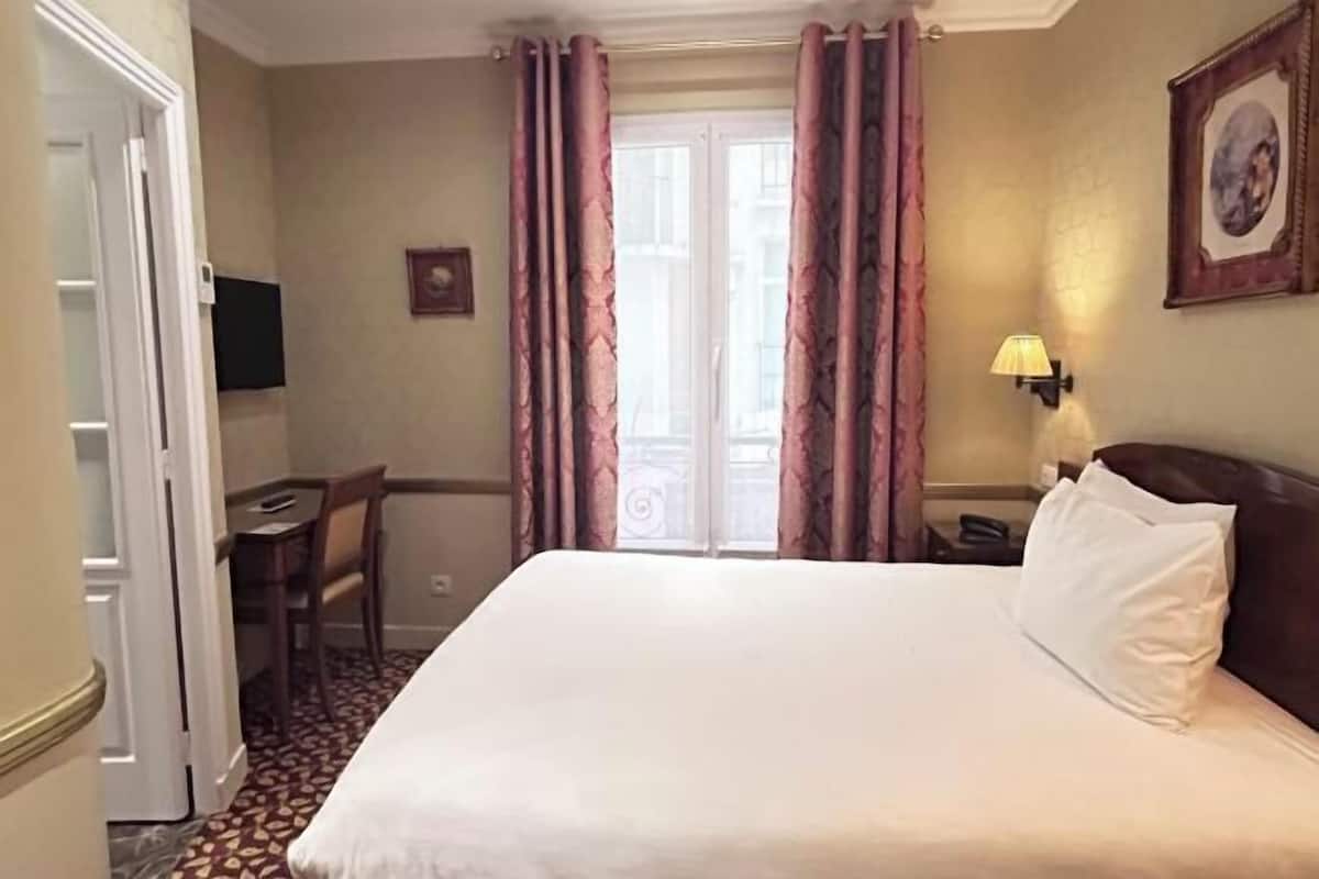 A hotel room with a bed, a desk, a chair, a window with curtains, and a wall-mounted lamp.