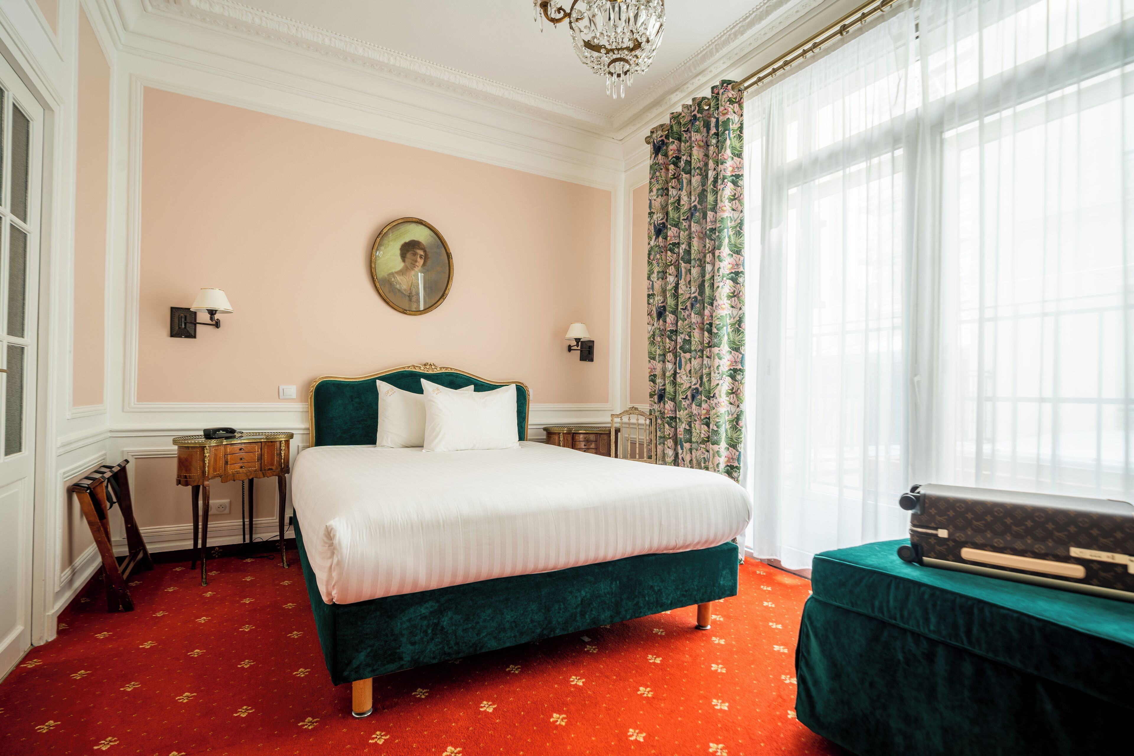 A hotel room with a large bed, a dresser, a chandelier, and a window with curtains.