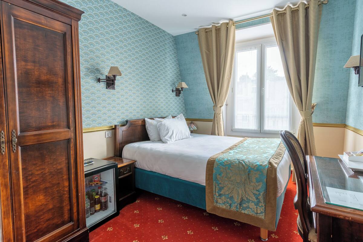 A hotel room with a bed, a desk, a wardrobe, and a small refrigerator.