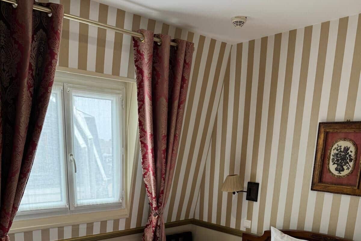 A hotel room with a large bed, striped wallpaper, a window with curtains, a bedside table, and a lamp.