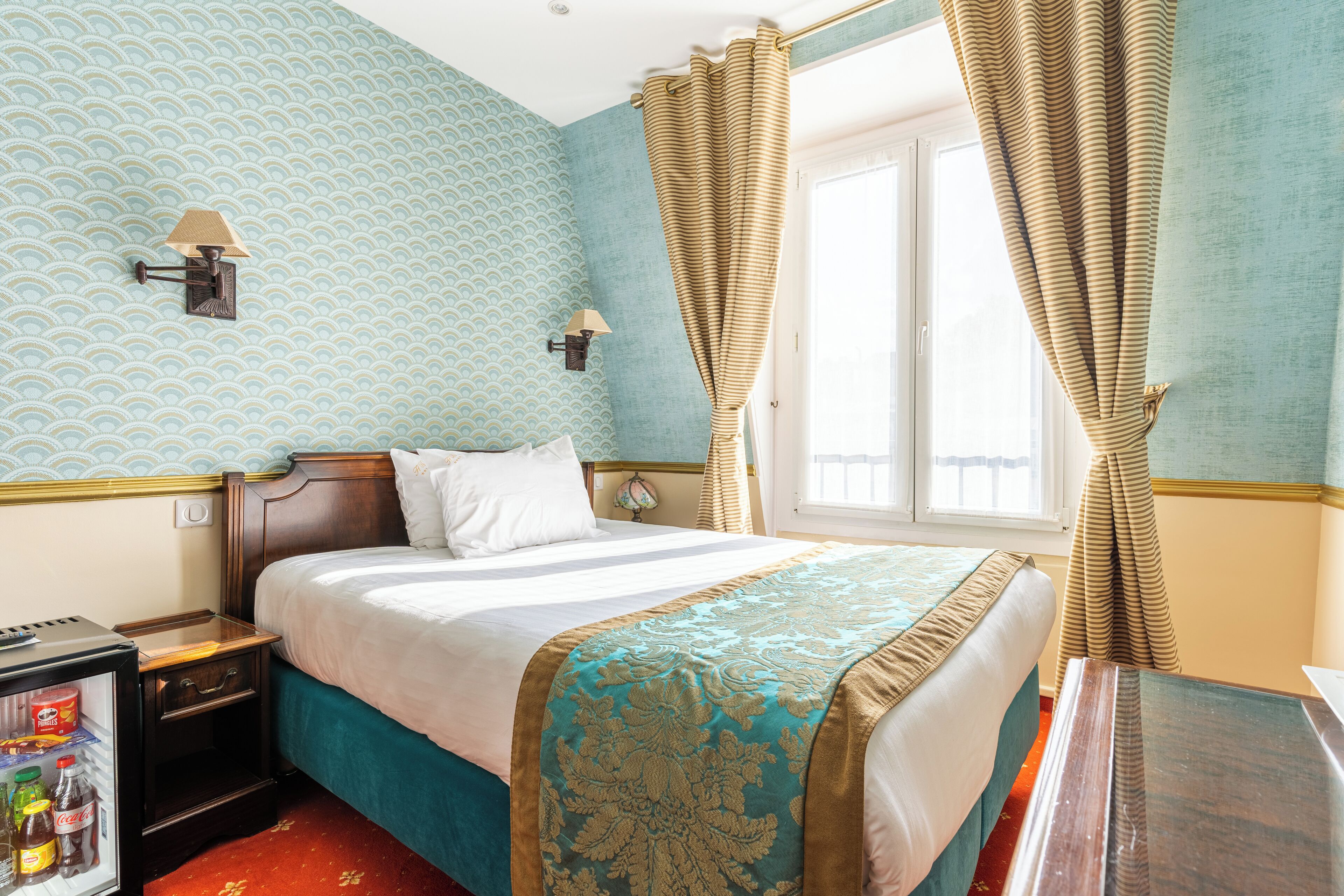 Une chambre d’hôtel comprenant un lit, une table de chevet avec une lampe, une fenêtre avec des rideaux et un mini-bar.