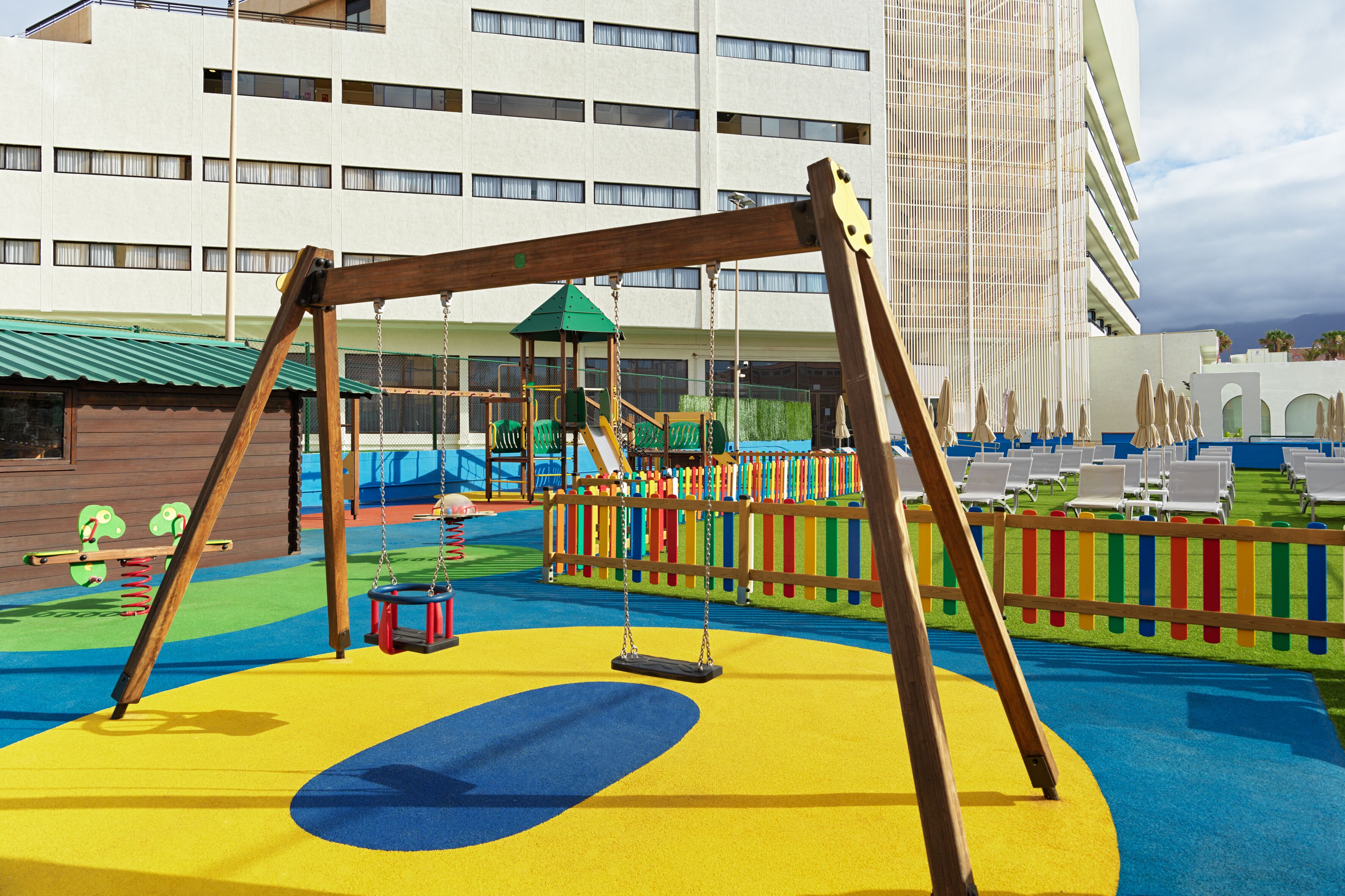 A playground with swings and slides, surrounded by a colorful fence, with a building in the background.