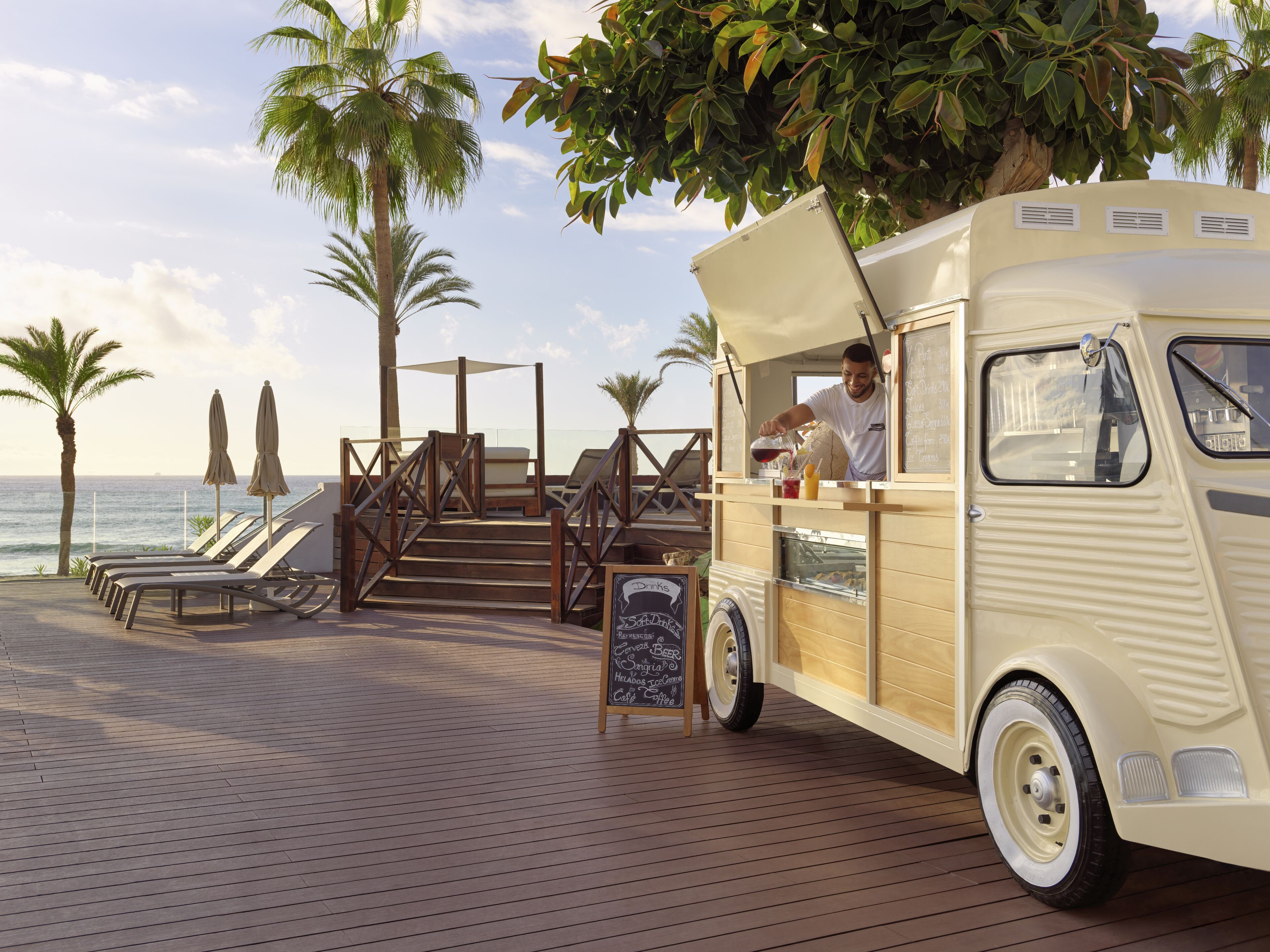 A vintage food truck with a chalkboard menu, located on a wooden deck with lounge chairs and palm trees, overlooking the ocean.