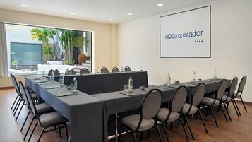 A conference room with a long table, chairs, and a projector screen displaying 'HIO Conquistador'.