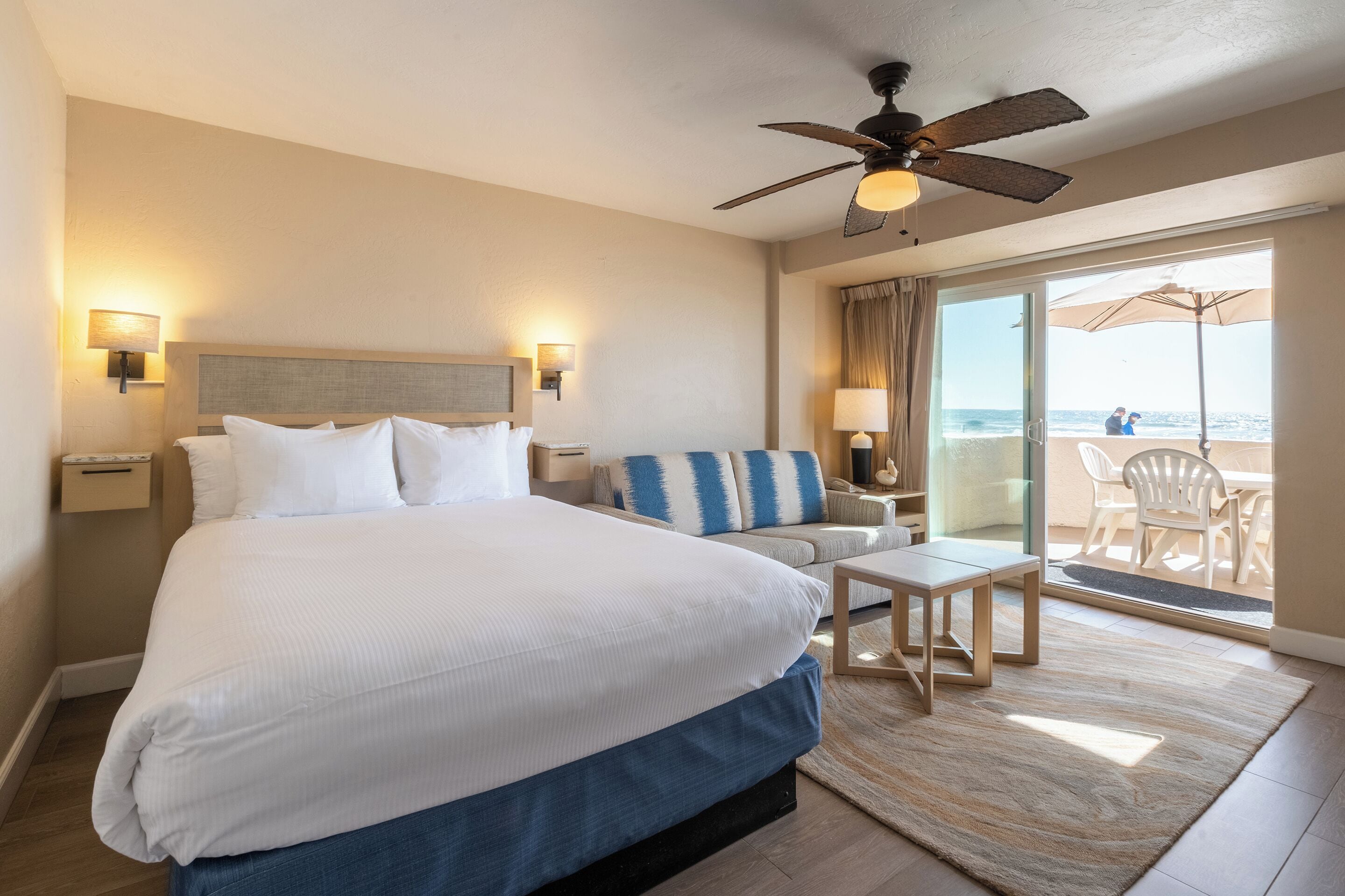A hotel room with a large bed, a sofa, a small table, and a balcony with a view of the beach.