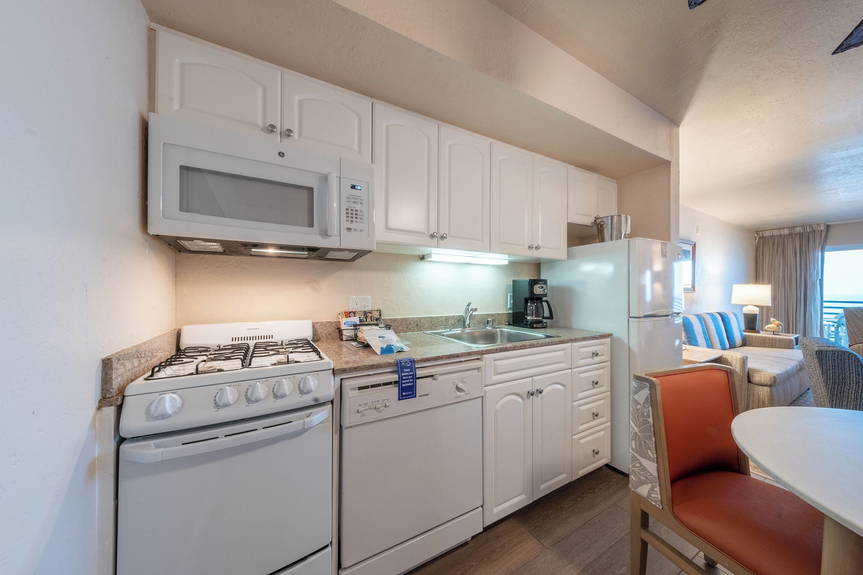Cocina compacta con electrodomésticos blancos, estufa a gas, microondas y lavavajillas. Hay una zona de comedor con una mesa y una silla naranja.