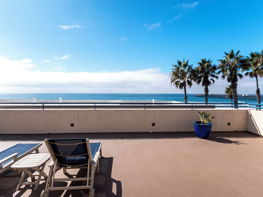 Terraço com cadeiras de descanso e uma mesa, com vista para o oceano, palmeiras e um píer ao longe.