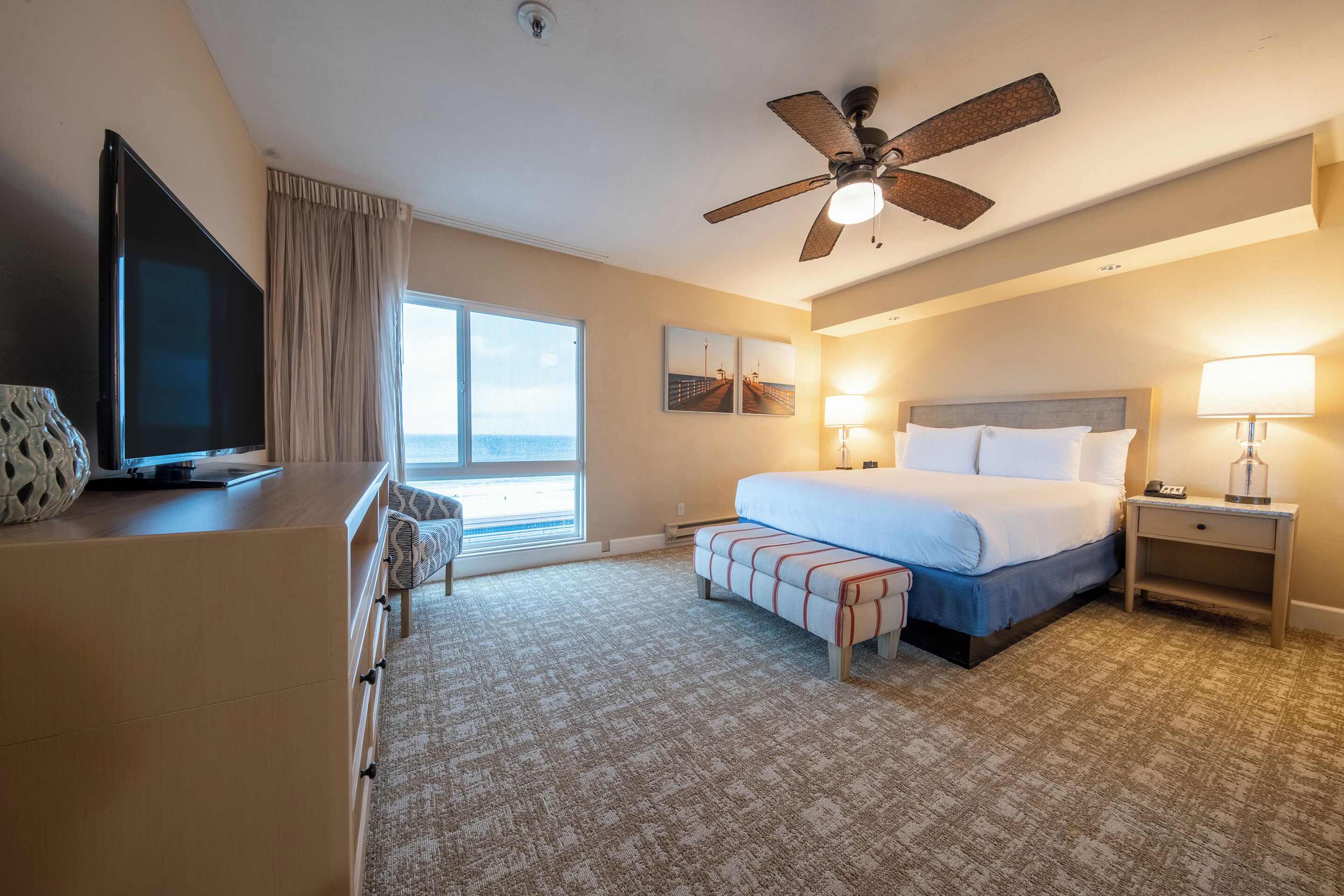 A hotel room with a bed, bedside lamps, a TV, a desk, and a view of the ocean.