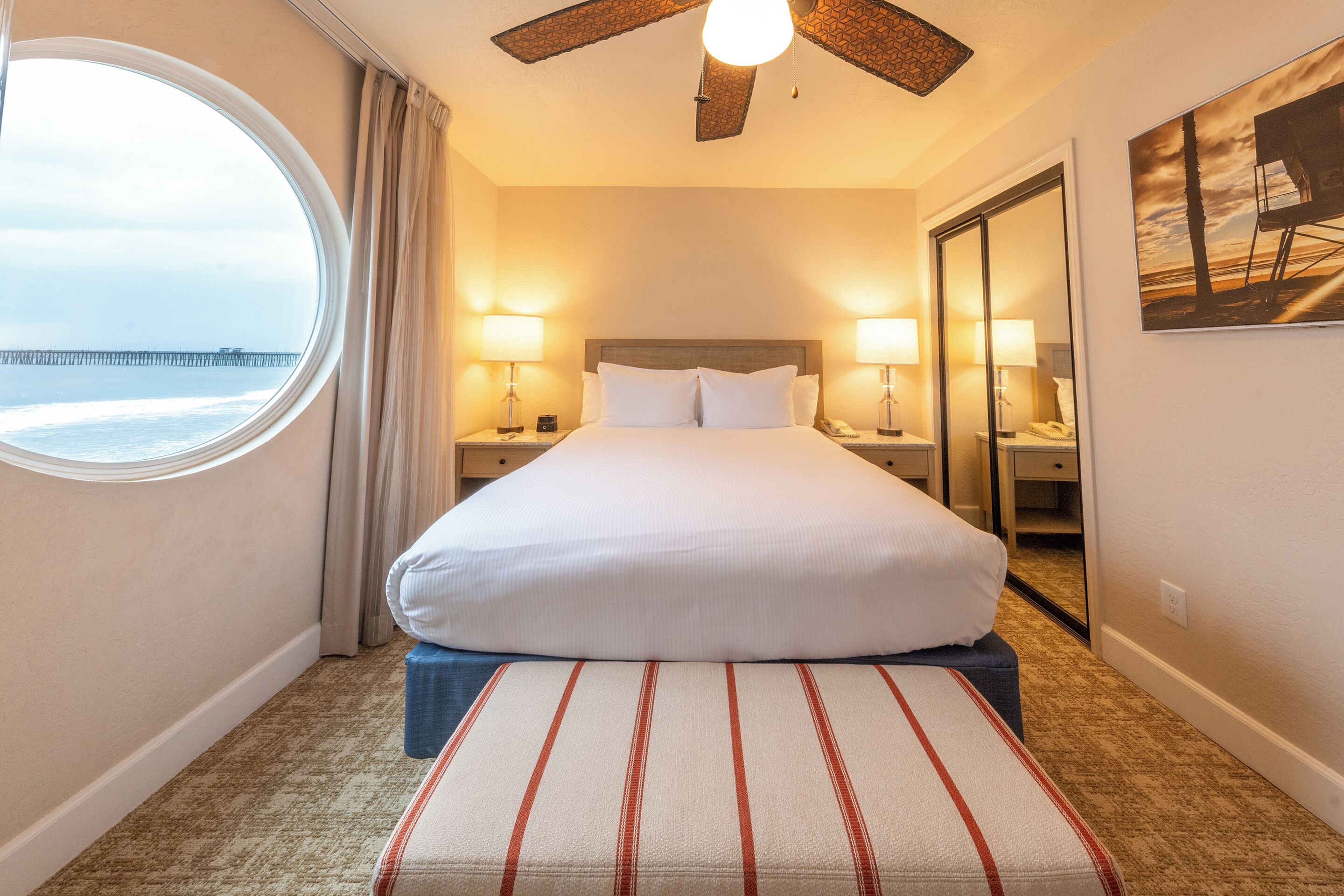 A hotel room with a large bed, two bedside lamps, a round window with a view of a pier, and a ceiling fan.