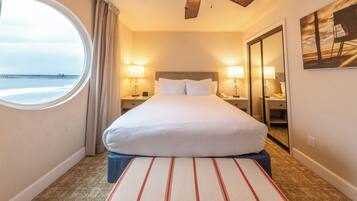 A hotel room with a large bed, two bedside lamps, a round window with a view of a pier, and a ceiling fan.