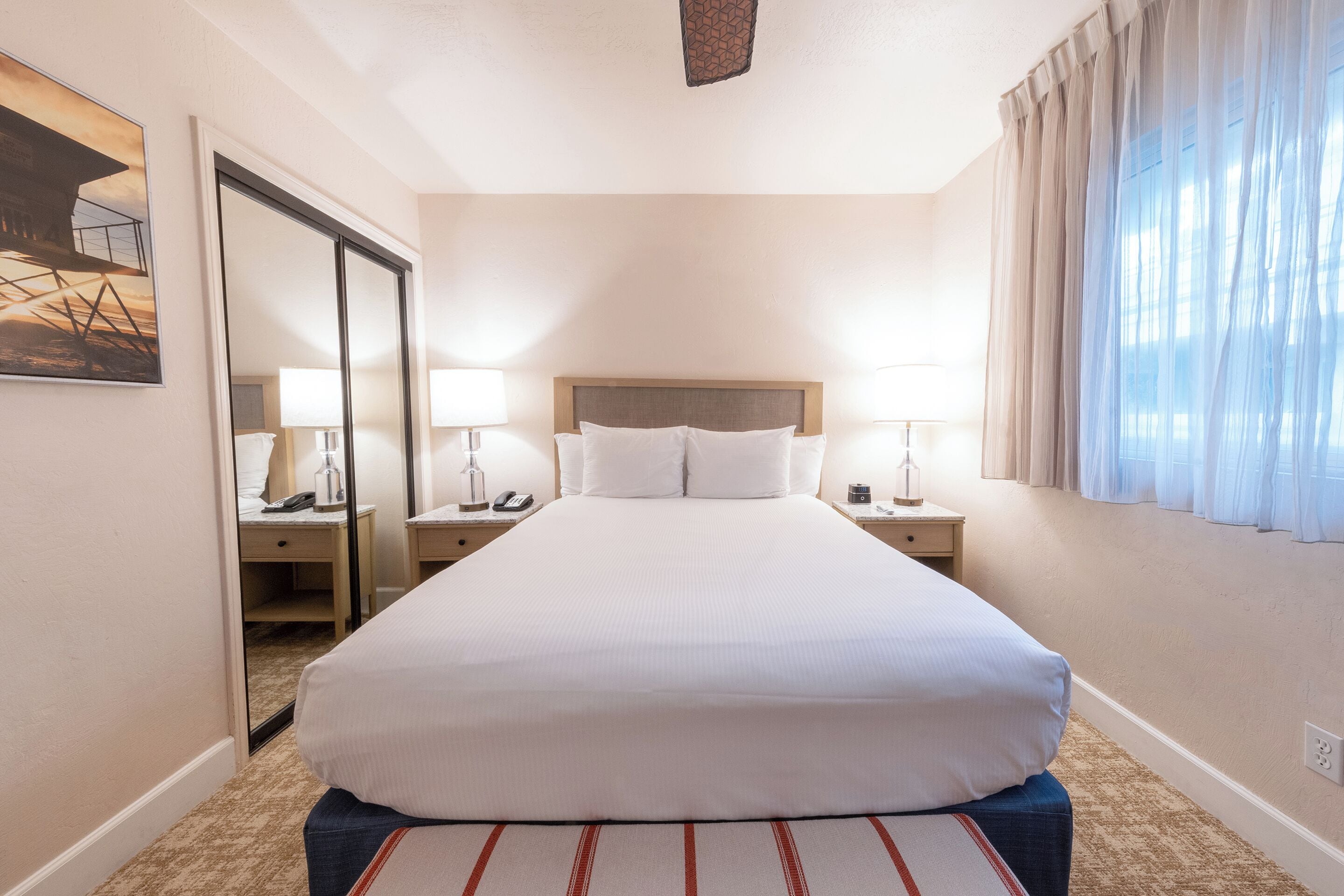 Una habitación de hotel con una cama grande, dos mesitas de noche con lámparas, un espejo y una ventana con cortinas.