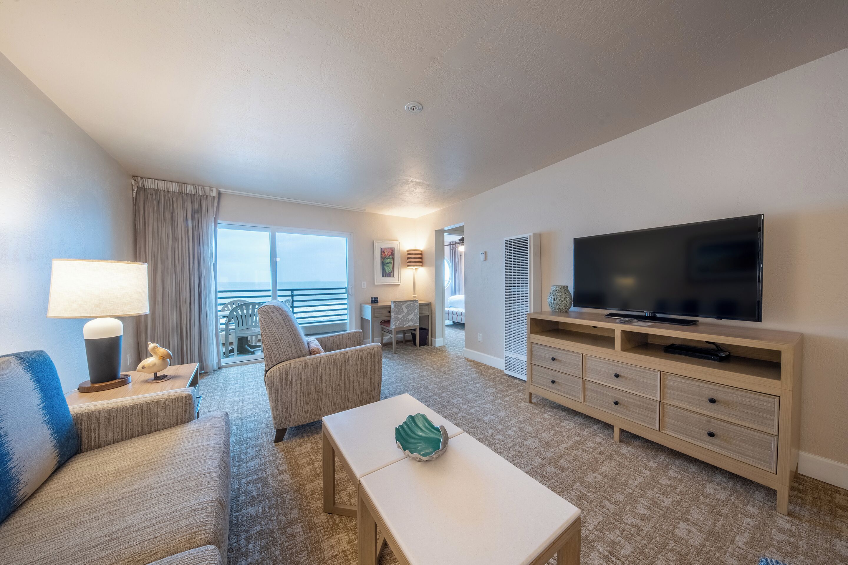 A hotel room with a large flat-screen TV, a sofa, a chair, a small table, and a balcony with a view.