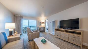 A hotel room with a large flat-screen TV, a sofa, a chair, a small table, and a balcony with a view.