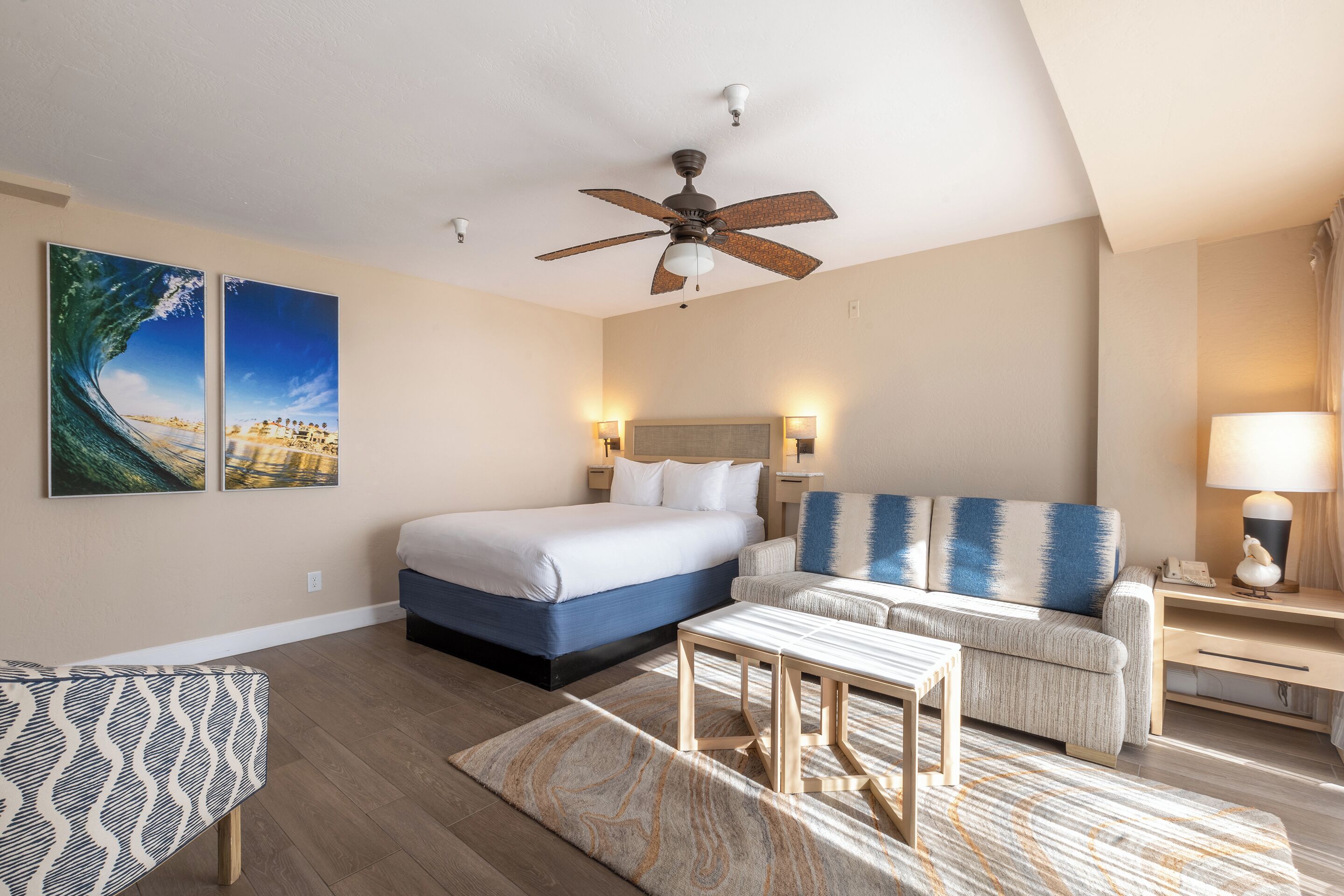 A hotel room with a bed, a sofa, a desk, a ceiling fan, and artwork on the walls.