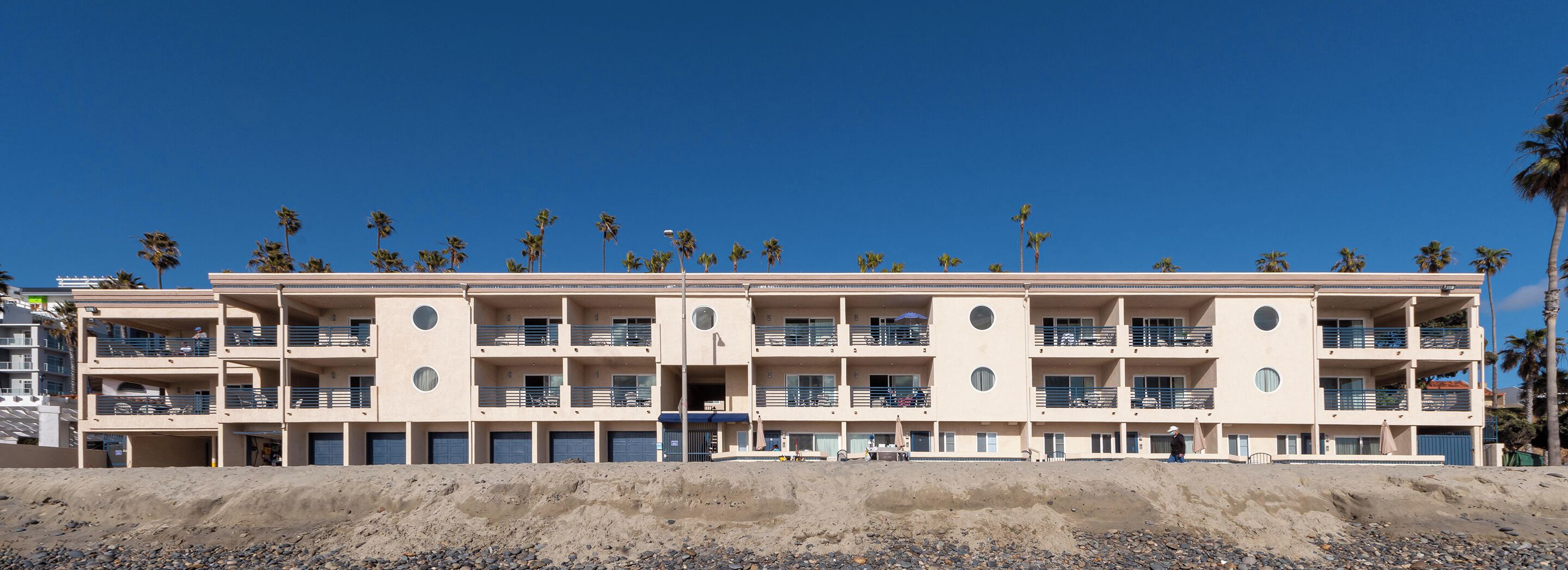 Un hotel de varios pisos con balcones, una playa arenosa al frente y palmeras en el fondo.