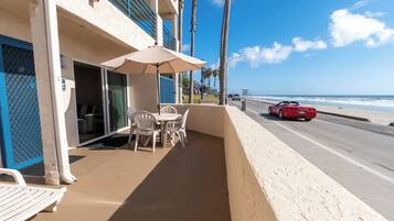 A balcony with a table and chairs, a red convertible on the road, and a view of the beach.