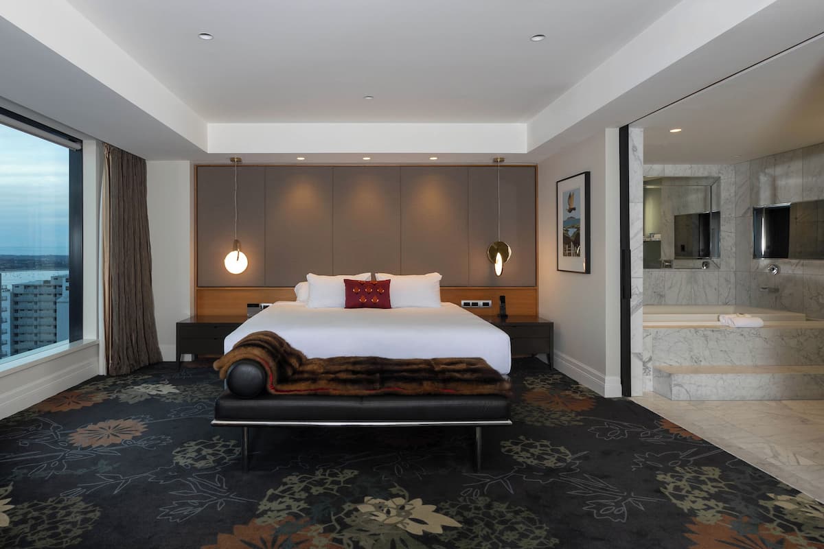 A modern hotel room with a large bed, a bench, a view of the city, and a bathroom with a marble floor.