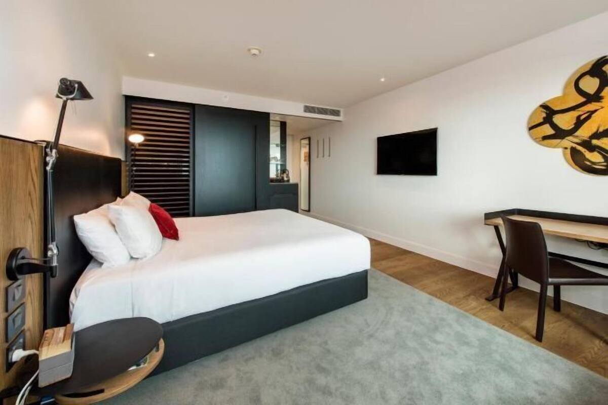 A modern hotel room with a bed, desk, chair, and a TV on the wall.