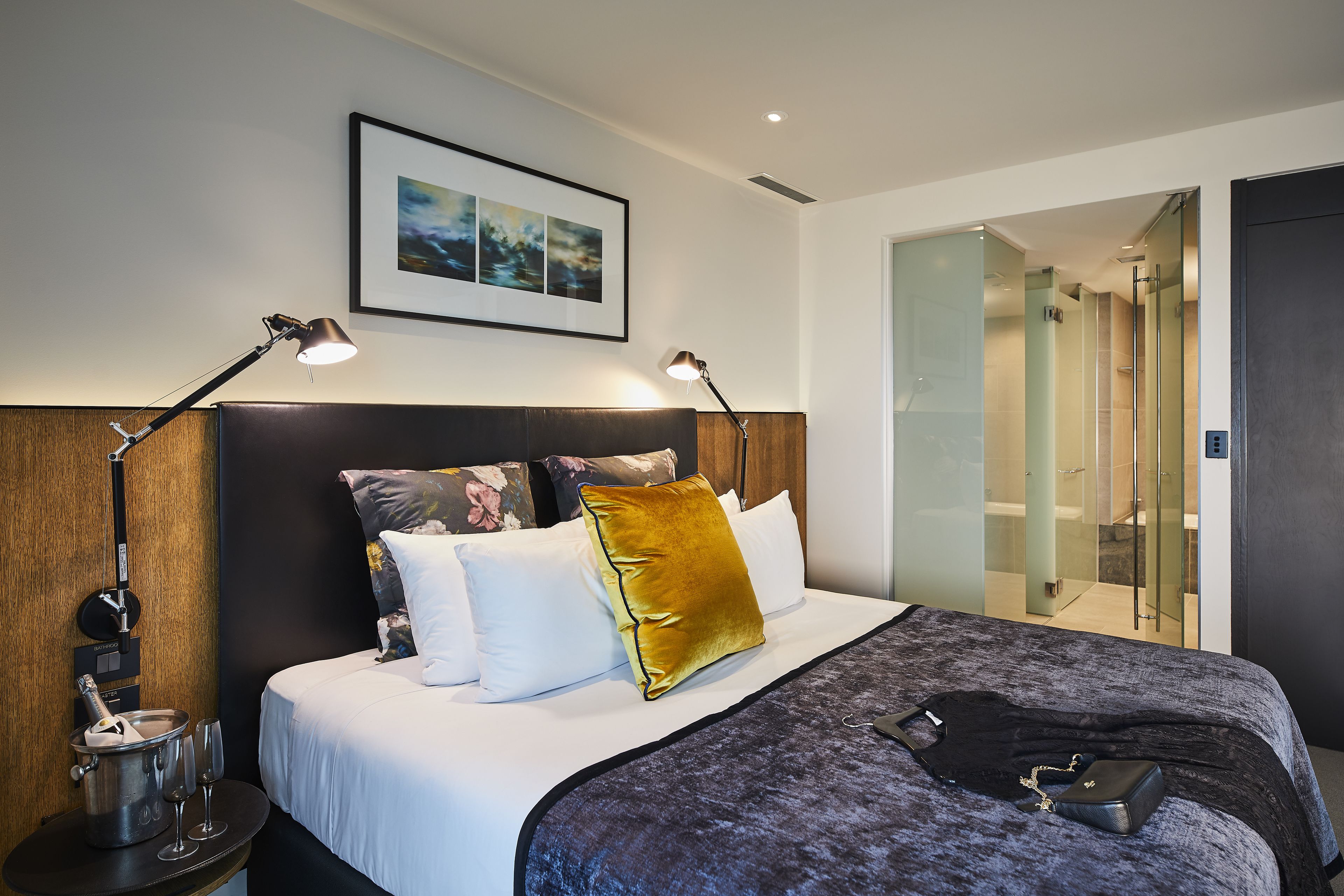 A modern hotel room with a large bed, two bedside lamps, a nightstand with a bottle and glasses, and a framed picture on the wall.