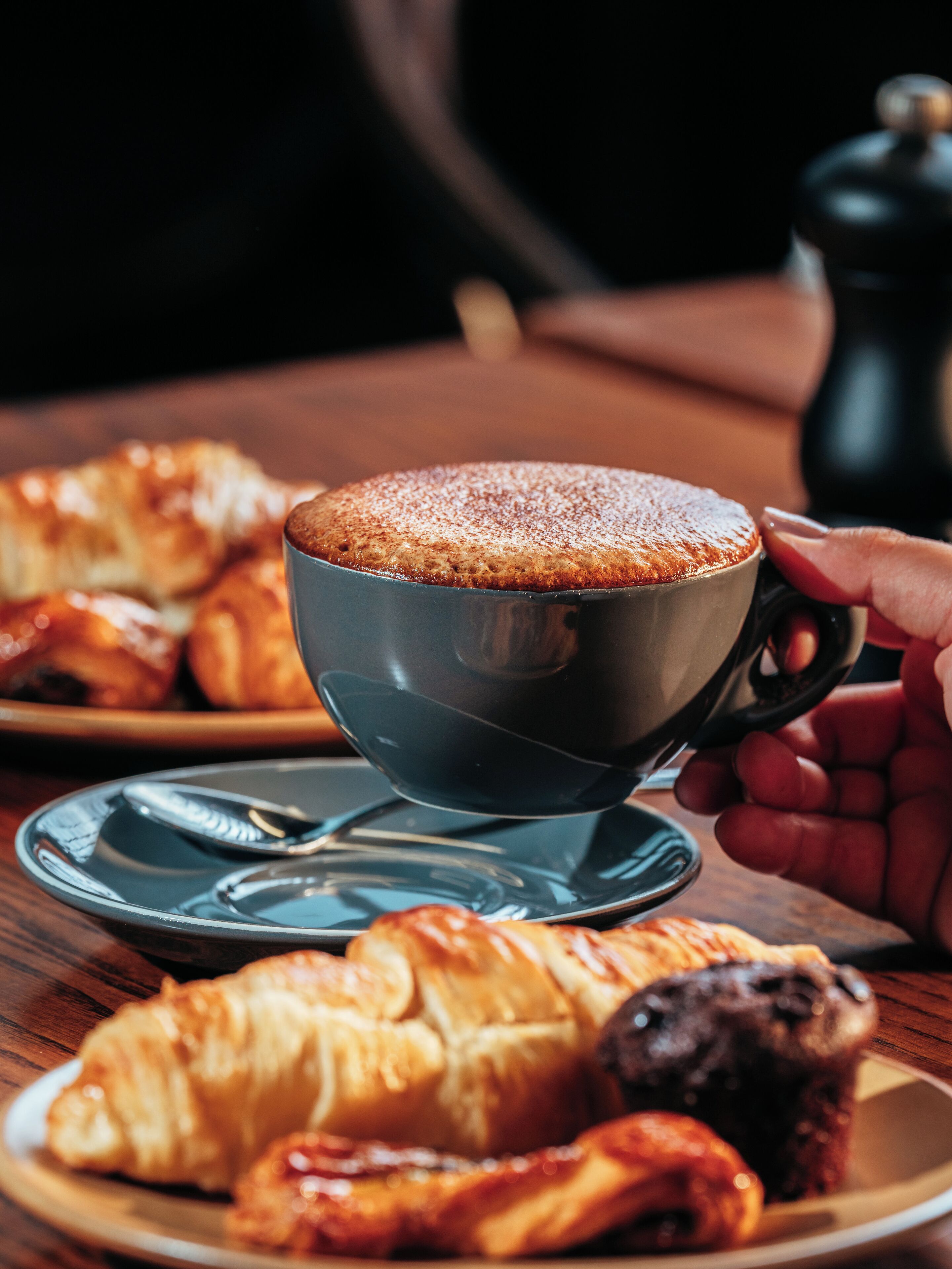 A cup of coffee with a saucer on a wooden table, accompanied by pastries and a pepper grinder.
