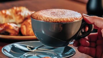 Uma chávena de café com pires sobre uma mesa de madeira, acompanhada por pastelaria e um moedor de pimenta.