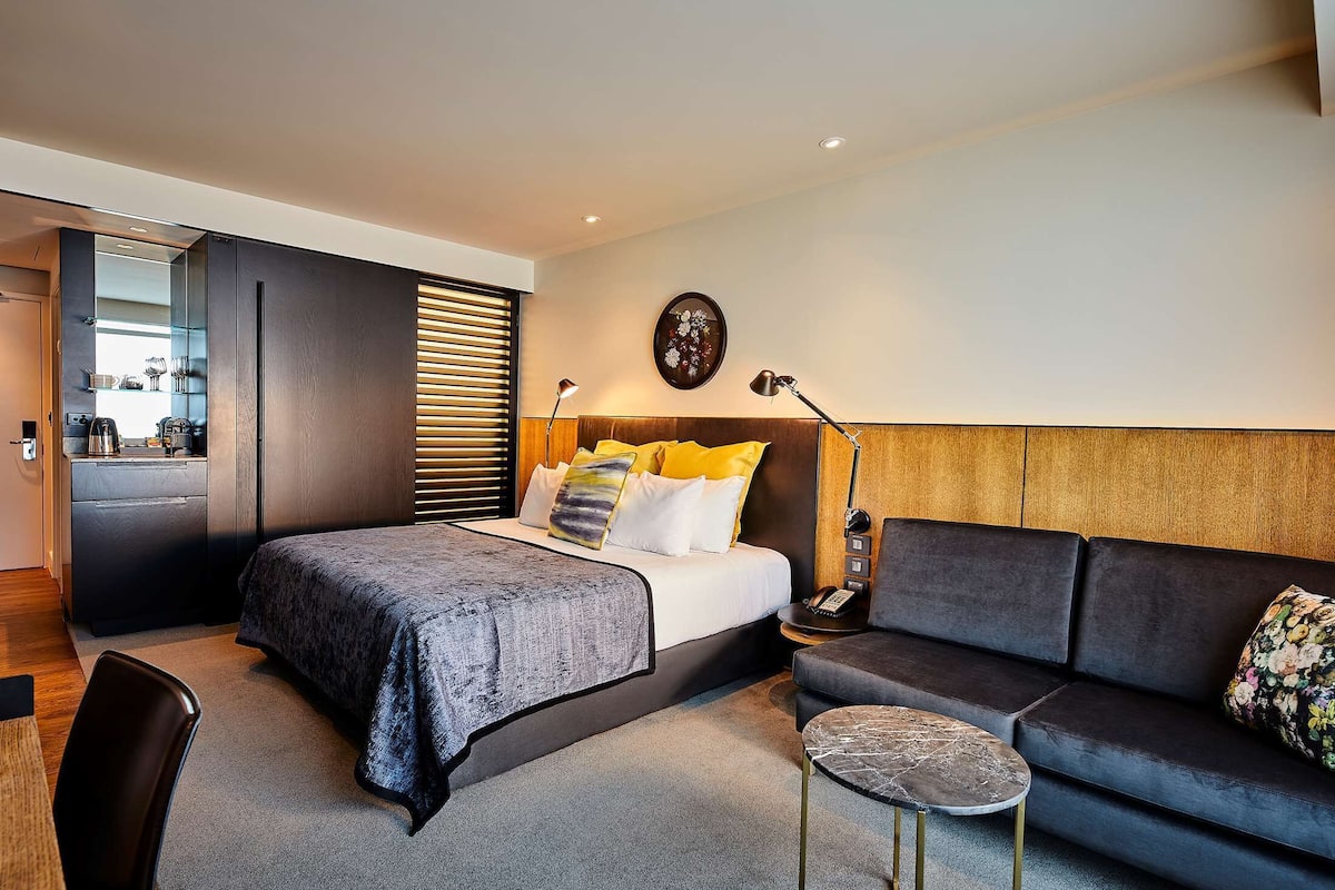 A modern hotel room with a bed, a sofa, a small table, and a desk with a lamp.