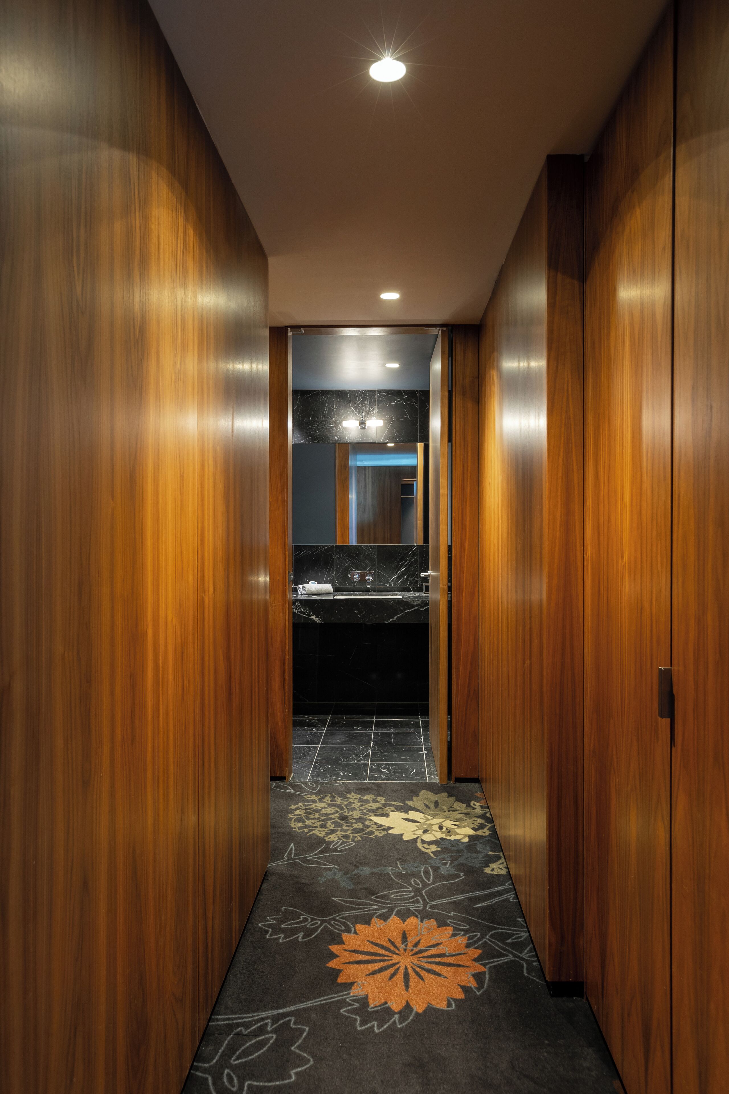 A hallway with wooden walls and a patterned carpet leading to a bathroom with a marble sink.
