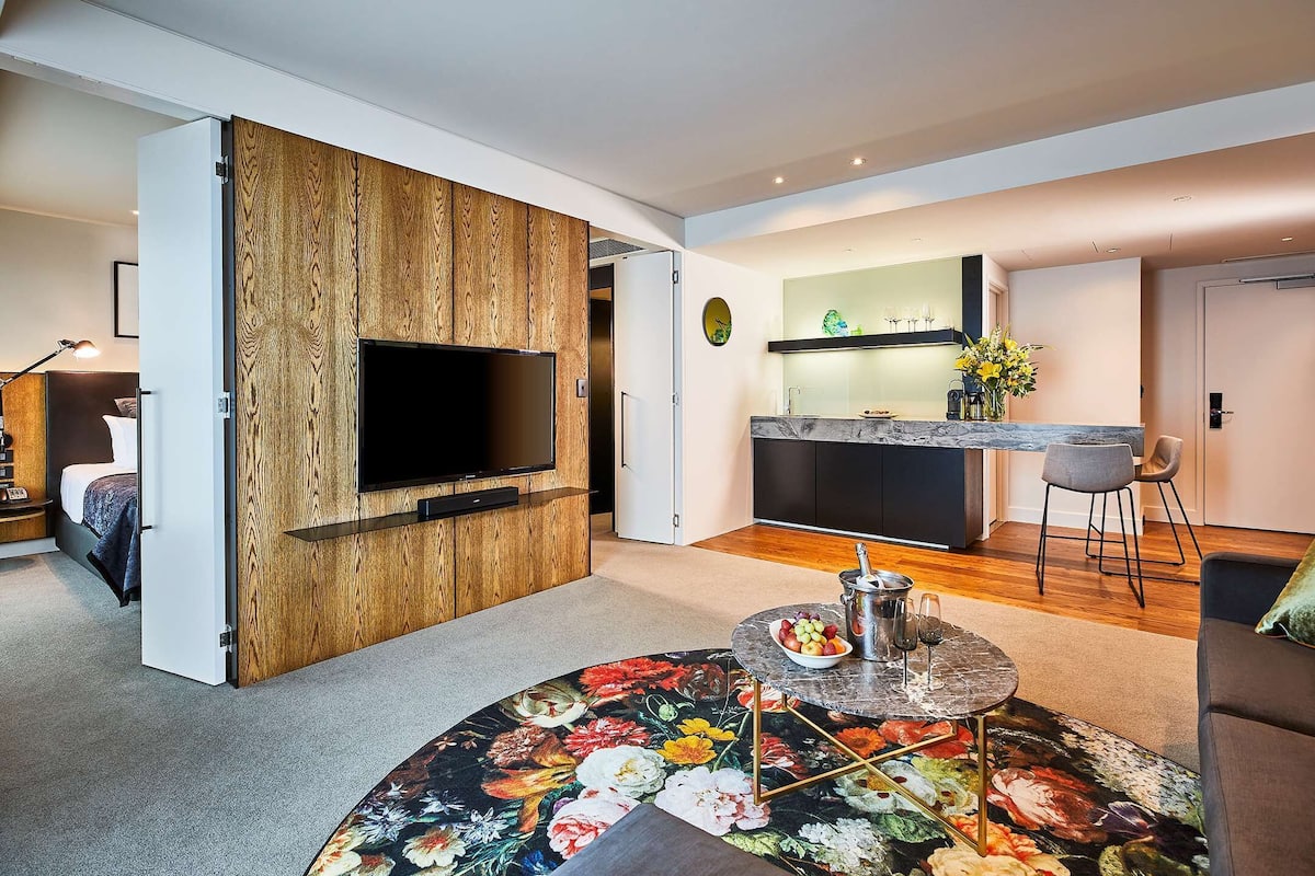A modern living room with a large TV, a dining area with a marble table, and a kitchen in the background.