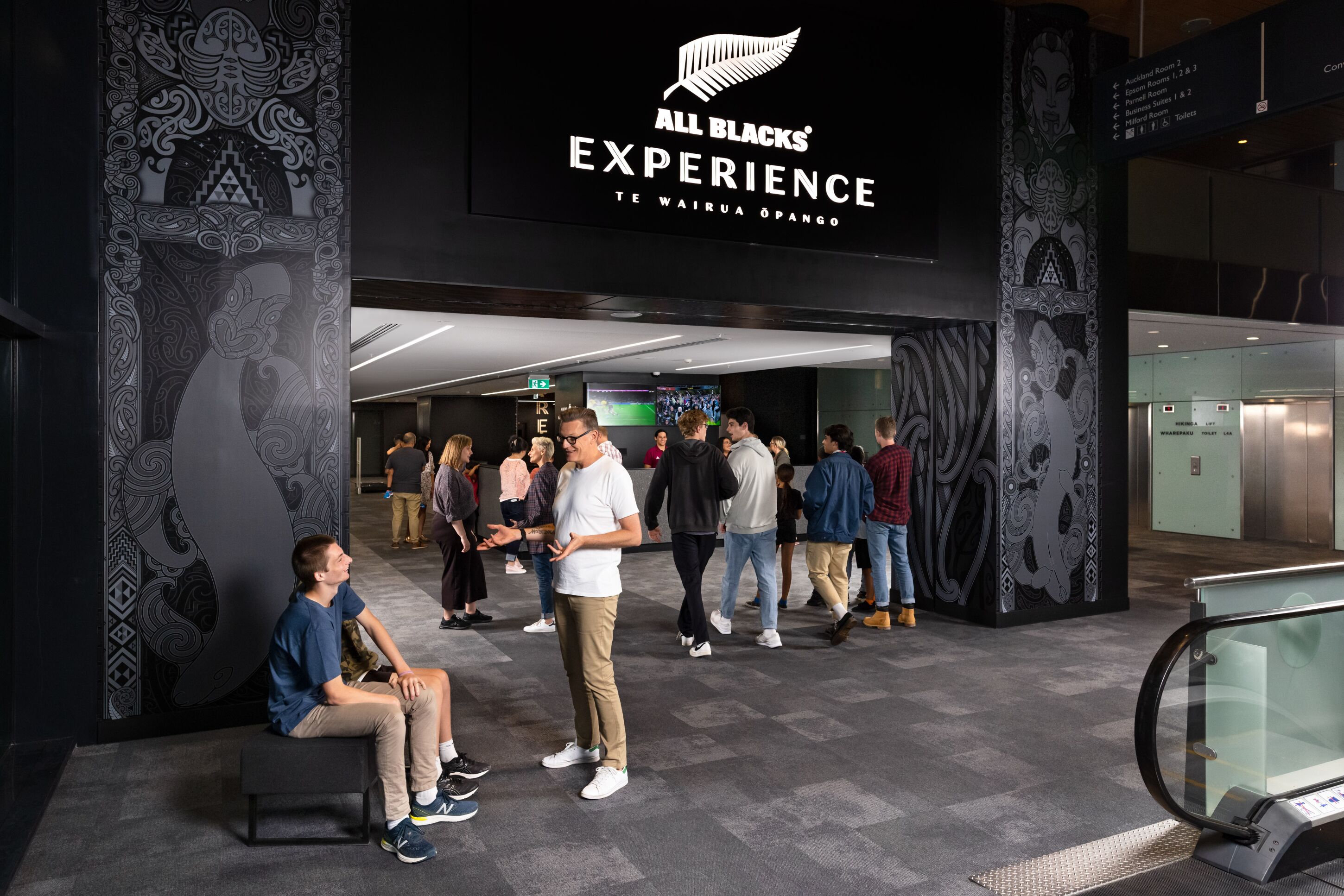A hotel entrance with a sign reading "All Blacks Experience" and a Maori phrase. People are gathered outside, some sitting and others standing.