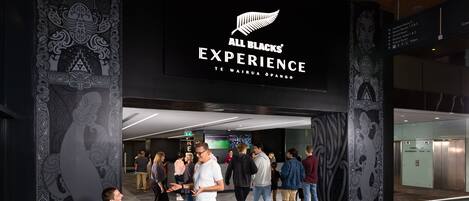 A hotel entrance with a sign reading "All Blacks Experience" and a Maori phrase. People are gathered outside, some sitting and others standing.